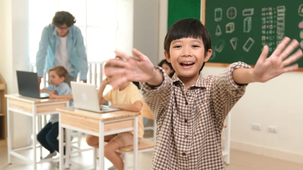 Asian boy looking at camera while waving hands and standing at blackboard. Skilled child greeting while diverse student study or learning about coding engineering prompt and generated AI. Pedagogy.