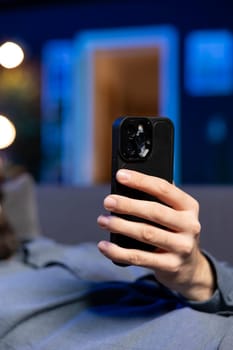 Focus on cellphone used by man in blurry background relaxing at home lounged on couch, texting friends. Close up shot of phone used by person laying down, chatting with mates online