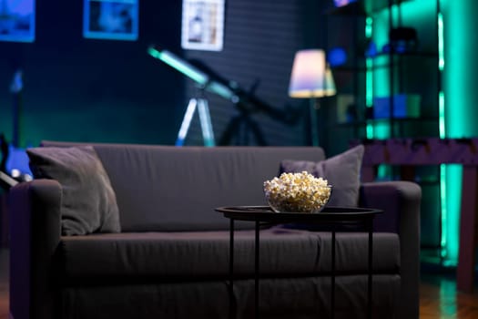 Bowl of popcorn on coffee table in front of cozy furniture in empty neon lit modern apartment. Snack prepared for movie night in dimly illuminated home theatre in front of couch