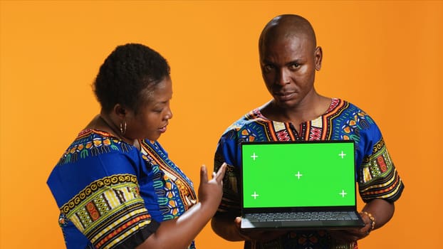 Ethnic couple pointing at laptop with greenscreen display, posing in studio with portable pc presenting isolated copyspace layout. Man and woman looking at blank mockup template on camera.