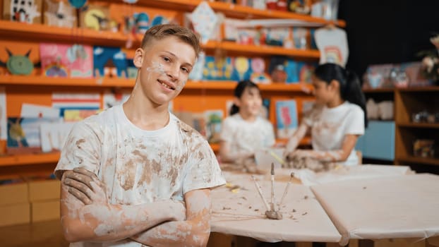 Smiling boy looking at camera and crossing arm with confident at workshop while diverse student having pottery class together. Happy caucasian student smile while pose with arm folded. Edification.