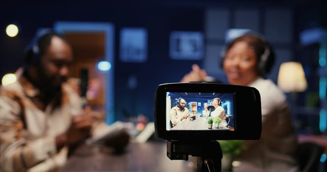 Focus on professional camera recording vlogger show host in blurry background interviewing guest in neon lights studio. Close up shot of equipment used to film online podcast episode