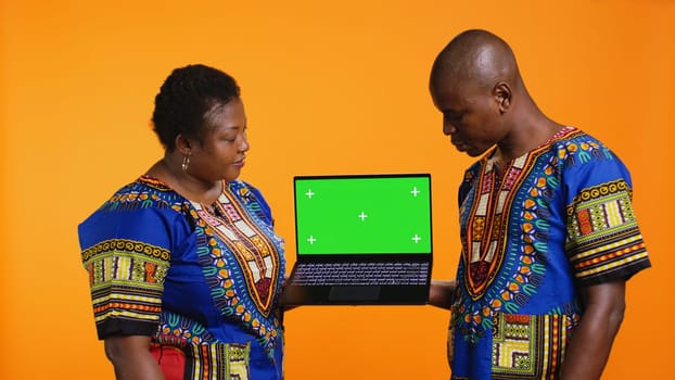 Ethnic people presenting laptop with greenscreen layout, showing blank mockup chromakey display and posing in studio. African american husband and wife pointing at isolated copyspace.