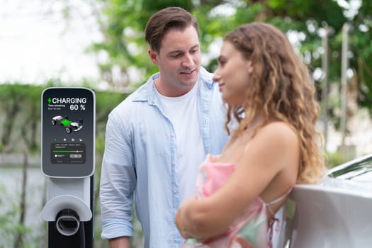 Happy and lovely couple with eco-friendly conscious recharging electric vehicle from EV charging station. EV car technology utilized as alternative transportation for future sustainability. Synchronos