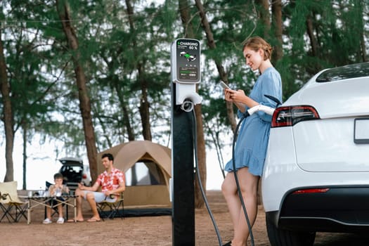 Outdoor adventure and family vacation camping at sea travel by eco friendly car. Woman or mother check car's battery with smartphone while charging EV car frin charging station in campsite. Perpetual