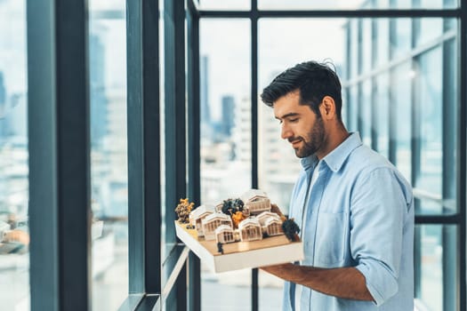 Businessman in casual outfit holding house model while checking house construction. Architect engineer inspect building model while standing near window with skyscraper. Civil engineering. Tracery.