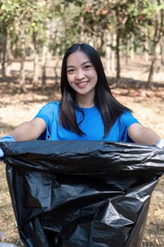 A cute young woman holds a garbage bag and a group of Asian volunteers collect garbage in plastic bags and clean up the area in the forest to preserve the natural ecosystem..