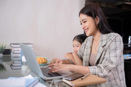 a single mother, sits at home working on a laptop with her daughter beside her watching and encouraging her..