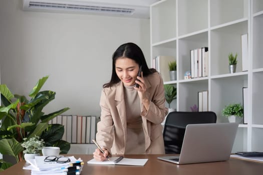 Young Asian business woman sits on the phone in an online business meeting using a laptop in a modern home office decorated with shady green plants..