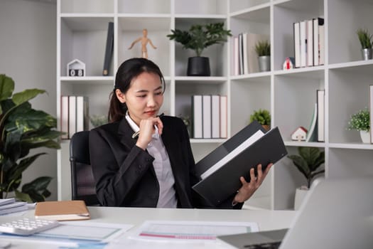 Young Asian business woman in a suit sits and reviews company work documents, checking information on a laptop on a work desk in a white office..