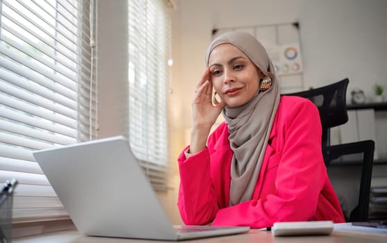 Young Muslim entrepreneur wearing hijab sits working with laptop managing personal business in private office..