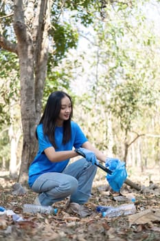 A cute young woman holds a garbage bag and a group of Asian volunteers collect garbage in plastic bags and clean up the area in the forest to preserve the natural ecosystem..