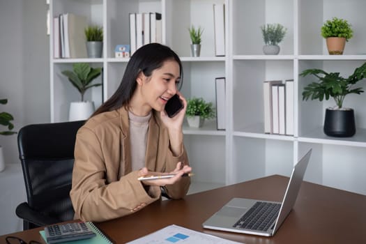 Young Asian business woman sits on the phone in an online business meeting using a laptop in a modern home office decorated with shady green plants..