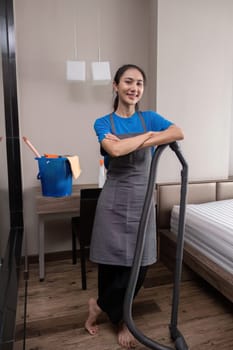A young female cleaner stands with a vacuum cleaner while vacuuming the bedroom..