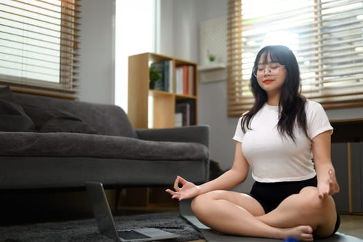 Peaceful young woman meditating in yoga lotus pose with closed eyes on mat at home.