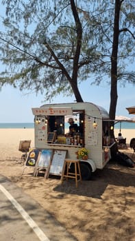 Rayong Thailand 13 March 2024, A vibrant food truck is parked on a sandy beach next to a lush tree, serving up delicious treats to beachgoers under the clear blue sky.
