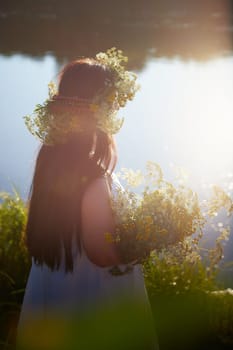 Adult mature brunette woman in a white dress, sundress and a wreath of flowers in summer by the water of river or lake in evening at sunset. Celebration of the Slavic pagan holiday of Ivan Kupala