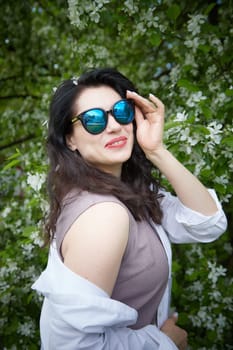 Funny brunette woman in sunglasses near Blossoms of apple tree in Spring Garden. Concept of face and body care. Scent of perfume and tenderness