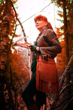 Mature model portraying a royal huntress with red curve hair is hunting with a crossbow in the in vibrant autumn forest in thematic photo shoot