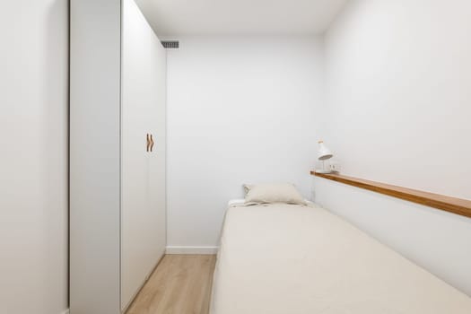 Horizontal shot of a single bed with a spacious white wardrobe, shelf and light fixture against white walls. Concept of an apartment bought with a mortgage for a single person.