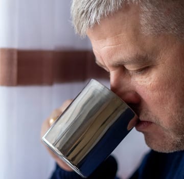 Portrait of the mid aged man with grey hair, wearing warm, dark blue sweater, drinking coffee.