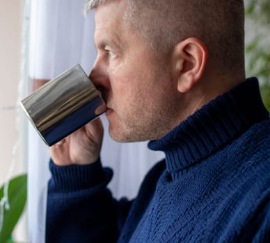 Portrait of the mid aged man with grey hair, wearing warm, dark blue sweater, drinking coffee.