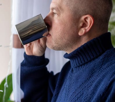 Portrait of the mid aged man with grey hair, wearing warm, dark blue sweater, drinking coffee.