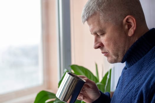 Portrait of the mid aged man with grey hair, wearing warm, dark blue sweater, drinking coffee.