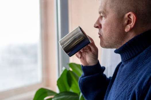 Portrait of the mid aged man with grey hair, wearing warm, dark blue sweater, drinking coffee.