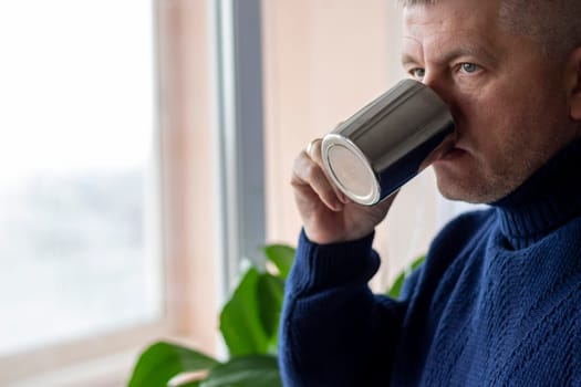 Portrait of the mid aged man with grey hair, wearing warm, dark blue sweater, drinking coffee.