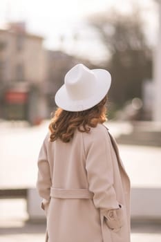 Back view of happy woman wearing hat and coat walking down street on sunny spring day. People, lifestyle, travel and vacations concept.