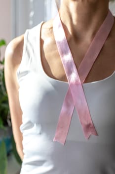 Shot of the woman in the white top against the white wall, with pink ribbon on her neck as a symbol of breast cancer awareness