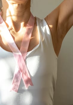 Shot of the woman in the white top against the white wall, with pink ribbon on her neck as a symbol of breast cancer awareness