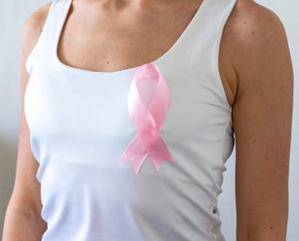 Shot of the woman against the white wall in the white top with pink ribbon, as a symbol of a breast cancer awareness