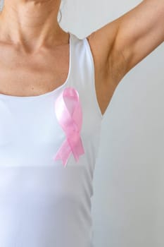 Shot of the woman against the white wall in the white top with pink ribbon, as a symbol of a breast cancer awareness