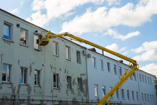 Worker in the basket of a car lift are repairing the facade of administrative building. Worker in hydraulic lifting ramp repair the window opening. Builder on lifting platform standing near the building wall, construction and repair works