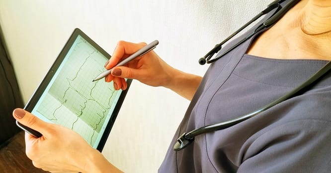 Shot of the female doctor in uniform against the white wall studying cardiogram on the tablet