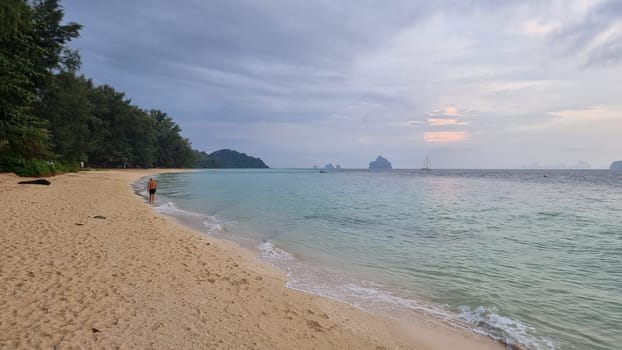A lone figure stands on a sandy beach, gazing out at the vast ocean with waves crashing gently on the shore. Koh Kradan Thailand