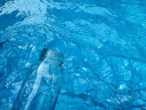 Clear glass bottle partially submerged in rippling blue water with reflections and refractions, top view. Clean water concept. High quality photo