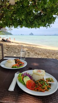 Two plates of delicious food are set on a table near the beach, with the ocean waves gently crashing in the background under the blue sky.Koh Kradan Thailand
