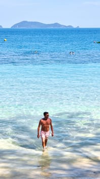 A man with a rugged appearance walks gracefully out of the glistening ocean water, the droplets sparkle in the sunlight as he emerges onto the sandy beach. Koh Wai Thailand