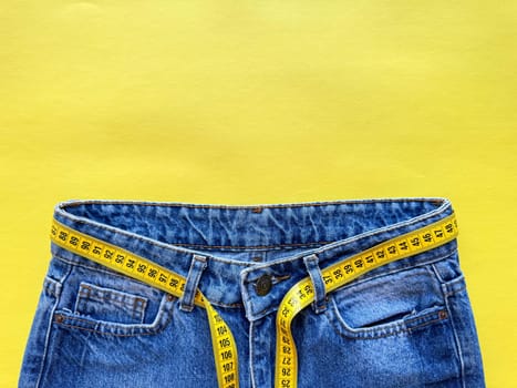 Blue jeans with measuring tape as belt on bright yellow background with copy space. Depicting weight loss, diet, and healthy lifestyle concept. Top view. High quality photo