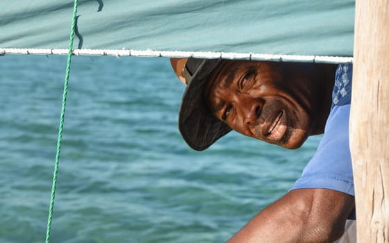 Anakao, Madagascar - May 03, 2019: Unknown Malagasy fisherman looking back under main green sail of his piroga (small fishing boat), green blue sea during sunny day in background