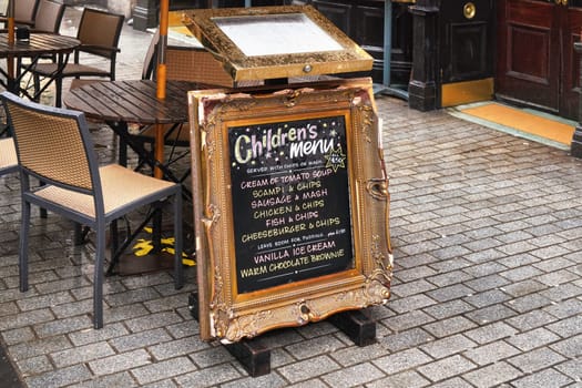 London, United Kingdom - February 02, 2019: Black Chalkboard with old ornate frame, and Children menu standing on pavement next to restaurant during wet day, chairs in background