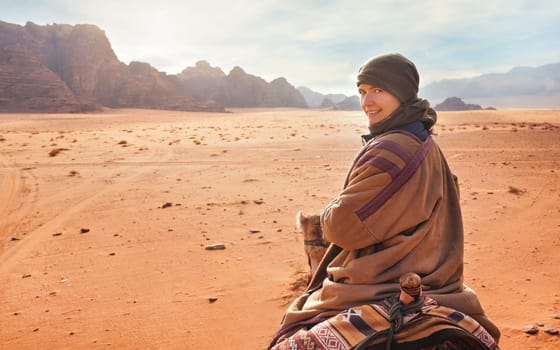 Young woman riding camel in desert, looking back over her shoulder, smiling. It's quite cold so she is wearing traditional Bedouin coat - bisht - and head scarf, mountains far background