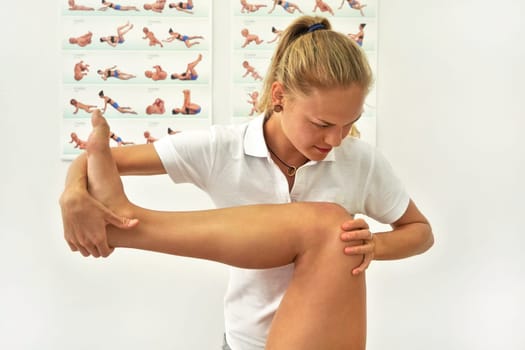 Young female physiotherapist exercising with her patient, flexing leg muscles while holding knee
