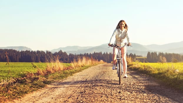 Young woman rides bicycle on dusty country road, afternoon sun shines at her, view from front