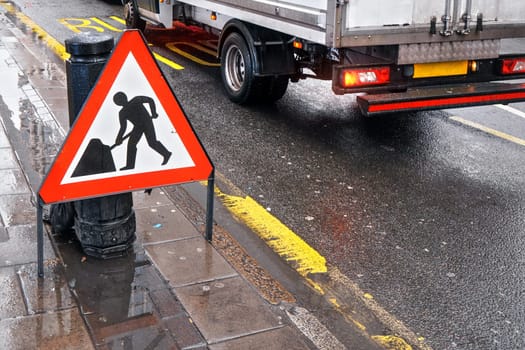 Warning roadworks sign at wet road next to pavement, van moving in background over Bus Stop writing on road