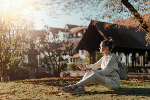 Young fashionable teenage girl with smartphone in park in autumn sitting at smiling. Trendy young woman in fall in park texting. Retouched, vibrant colors. Beautiful blonde teenage girl wearing casual modern autumn outfit sitting in park in autumn. Retouched, vibrant colors, brownish tones.