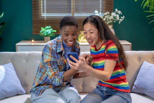 Black and asian friends are sharing a moment of leisure on a couch by a window, smiling at a cell phone excited and surprised. They look happy and comfortable, with a houseplant in the corner of the room. High quality photo